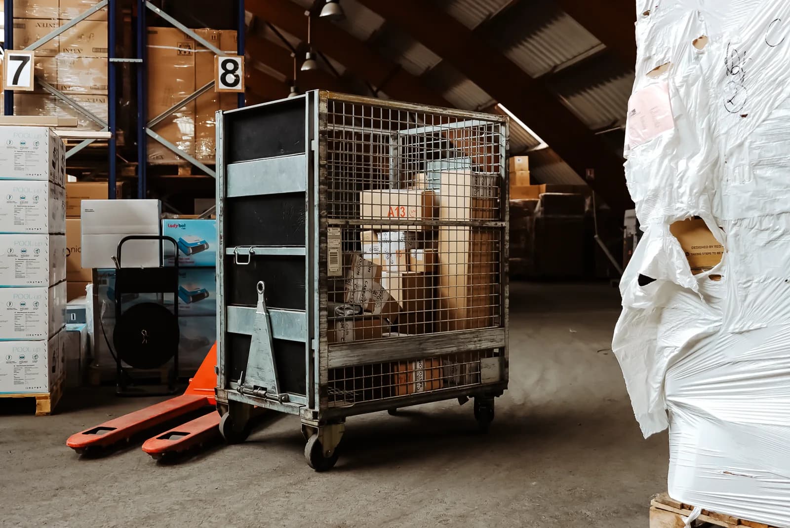 A wire cage cart loaded with boxes next to a pallet jack, in front of numbered racks in the Cabesto warehouse.