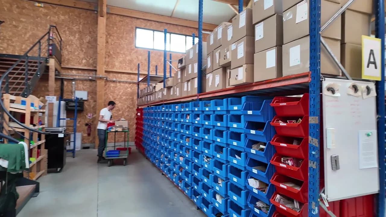 Wide view down a picking aisle at La Ferme de Sainte-Marthe, with location bins and an operator beside a cart.
