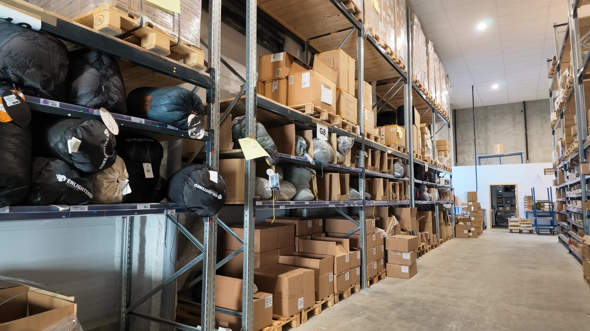 A warehouse aisle: sleeping bags on the shelves, with cartons and overstock pallets on the upper levels.